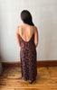 A woman viewed from behind wearing a dark brown floral lace slip dress with thin spaghetti straps and a low open back, standing on a wooden floor against a plain off-white wall.