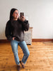A woman takes a mirror selfie indoors, wearing a dark brown knit sweater, blue jeans, and brown loafers, standing on a wooden floor in front of a white wall.