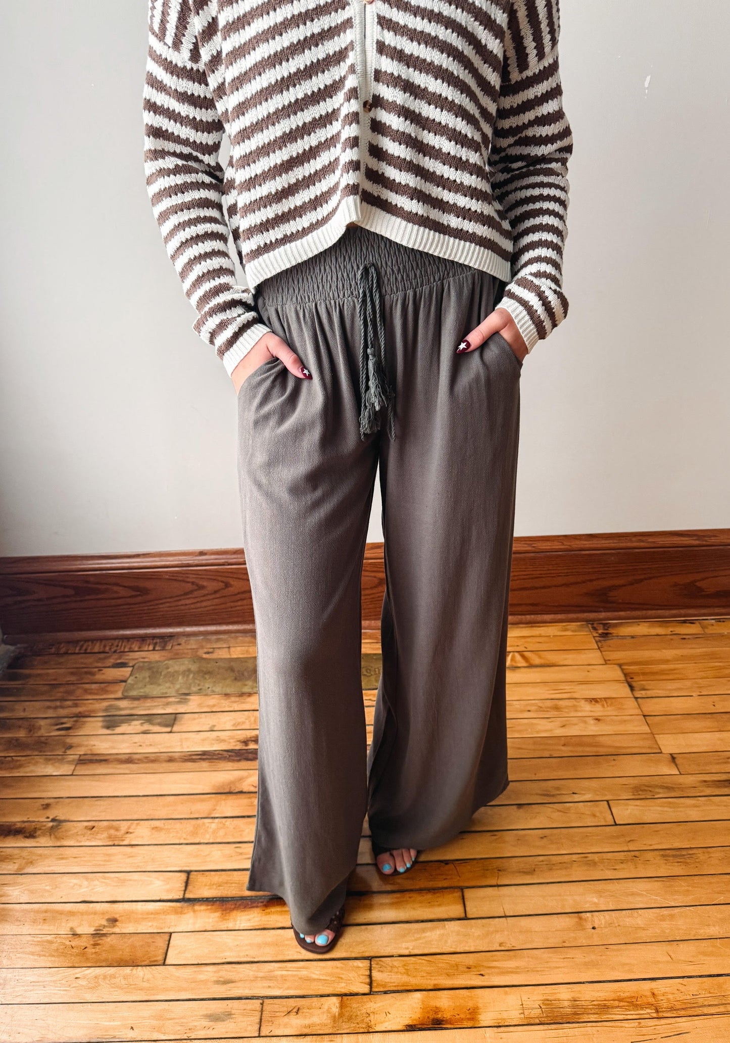 A person wearing loose, wide-leg taupe pants with a drawstring waist and side pockets, paired with a brown and cream striped cardigan, standing on a wooden floor against a plain wall.