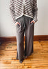 A person wearing loose, wide-leg taupe pants with a drawstring waist and side pockets, paired with a brown and cream striped cardigan, standing on a wooden floor against a plain wall.