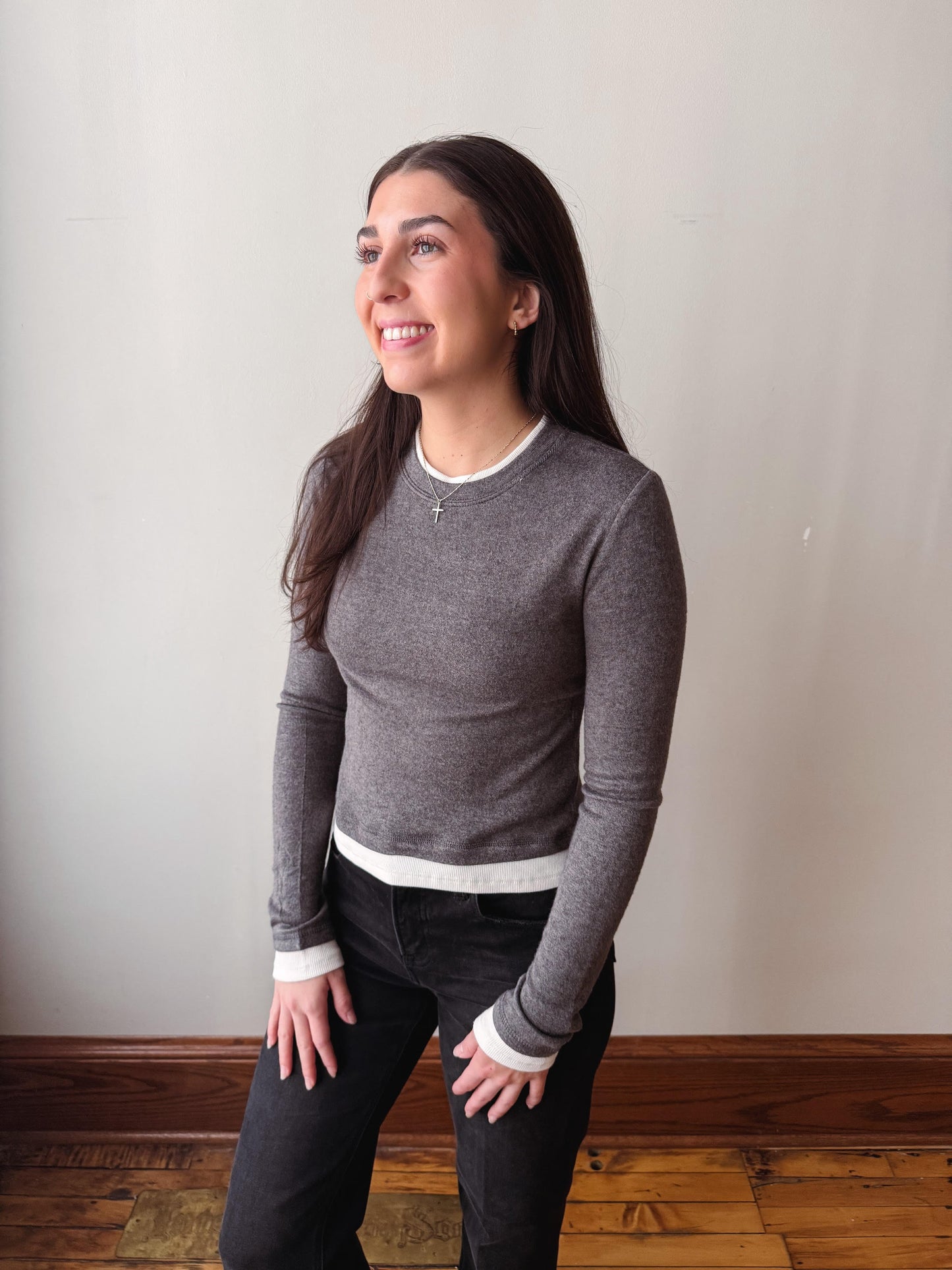 A woman wearing a heather gray long-sleeve crewneck sweater with white ribbed trim at the collar, hem, and cuffs, paired with black pants, standing against a plain off-white wall.