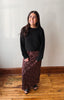 A woman stands indoors wearing a black long-sleeve knit top and a dark brown patterned maxi skirt, paired with black sandals, against a plain off-white wall and wooden floor.