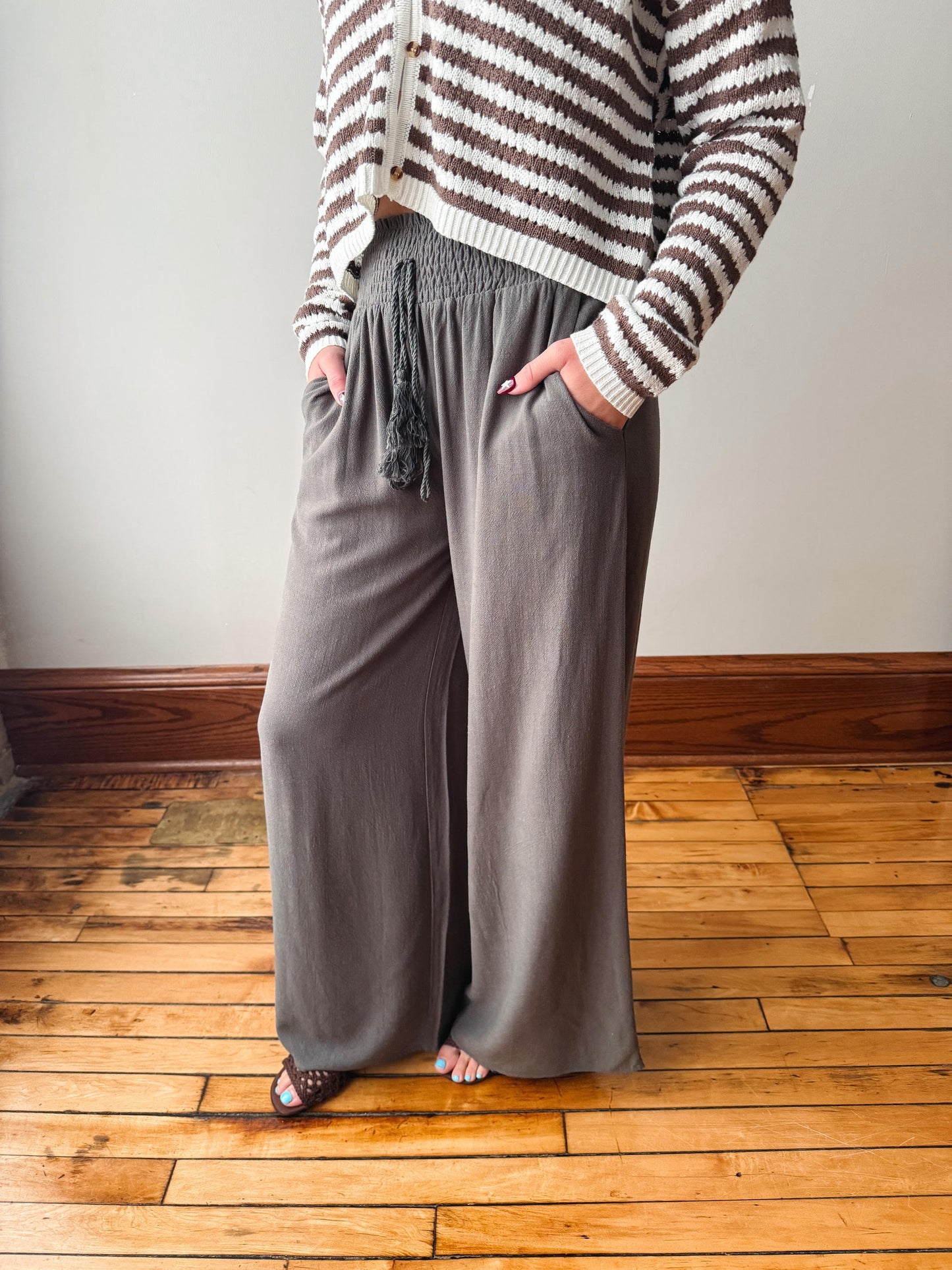 A person wearing wide-leg taupe gray pants with a drawstring waist, paired with a brown and cream striped cardigan, standing on a wooden floor against a neutral wall.