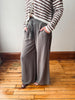 A person wearing wide-leg taupe gray pants with a drawstring waist, paired with a brown and cream striped cardigan, standing on a wooden floor against a neutral wall.