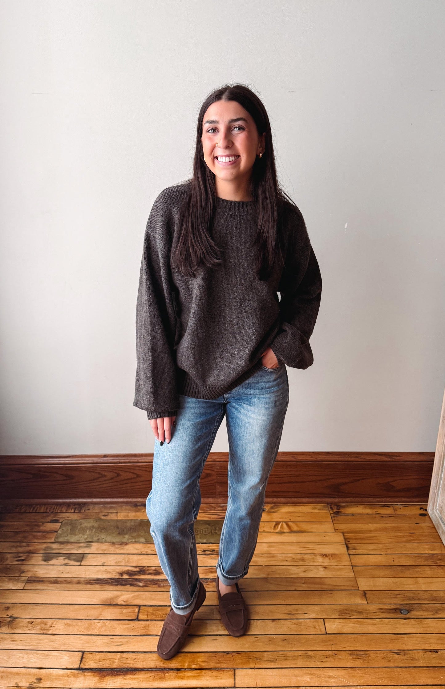 A woman stands in a room with a light gray wall and wooden floor, wearing a dark brown knit sweater, blue jeans, and brown suede loafers.