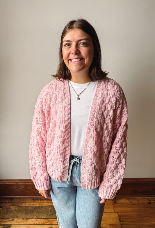 A smiling woman wearing a chunky pink cable-knit cardigan over a white t-shirt and light blue jeans, standing against a neutral beige wall with a wooden floor.