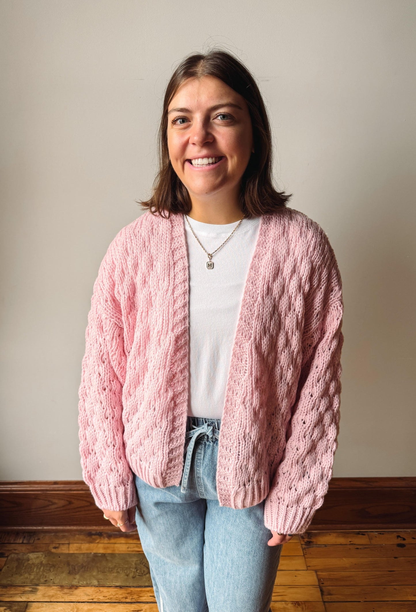 A smiling woman wearing a chunky pink cable-knit cardigan over a white t-shirt and light blue jeans, standing against a neutral beige wall with a wooden floor.