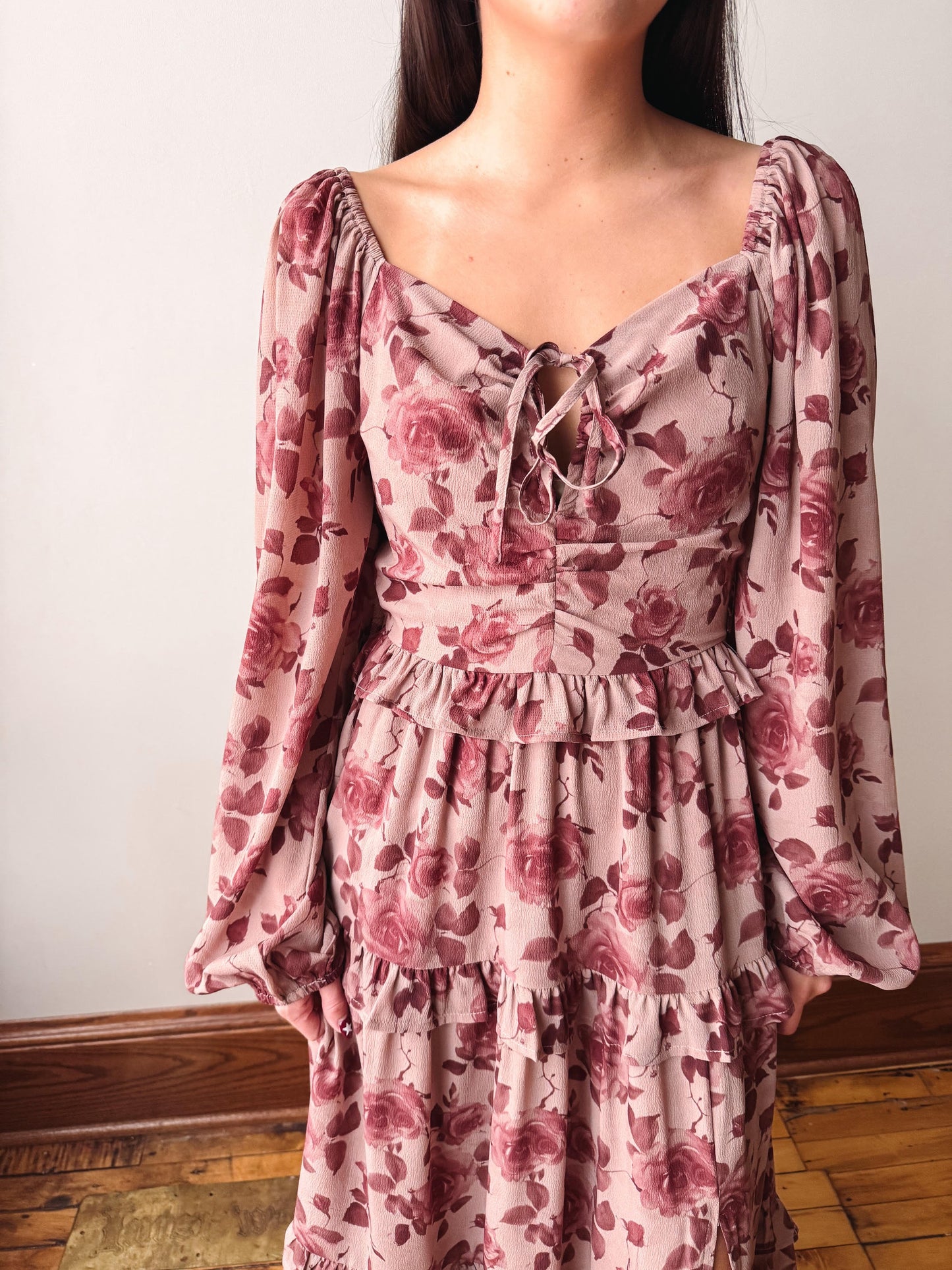 A woman wearing a dusty rose pink long-sleeve tiered dress with a dark red rose floral print, featuring a tie-neck detail and ruffled waist, standing against a plain off-white wall.