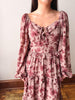 A woman wearing a dusty rose pink long-sleeve tiered dress with a dark red rose floral print, featuring a tie-neck detail and ruffled waist, standing against a plain off-white wall.