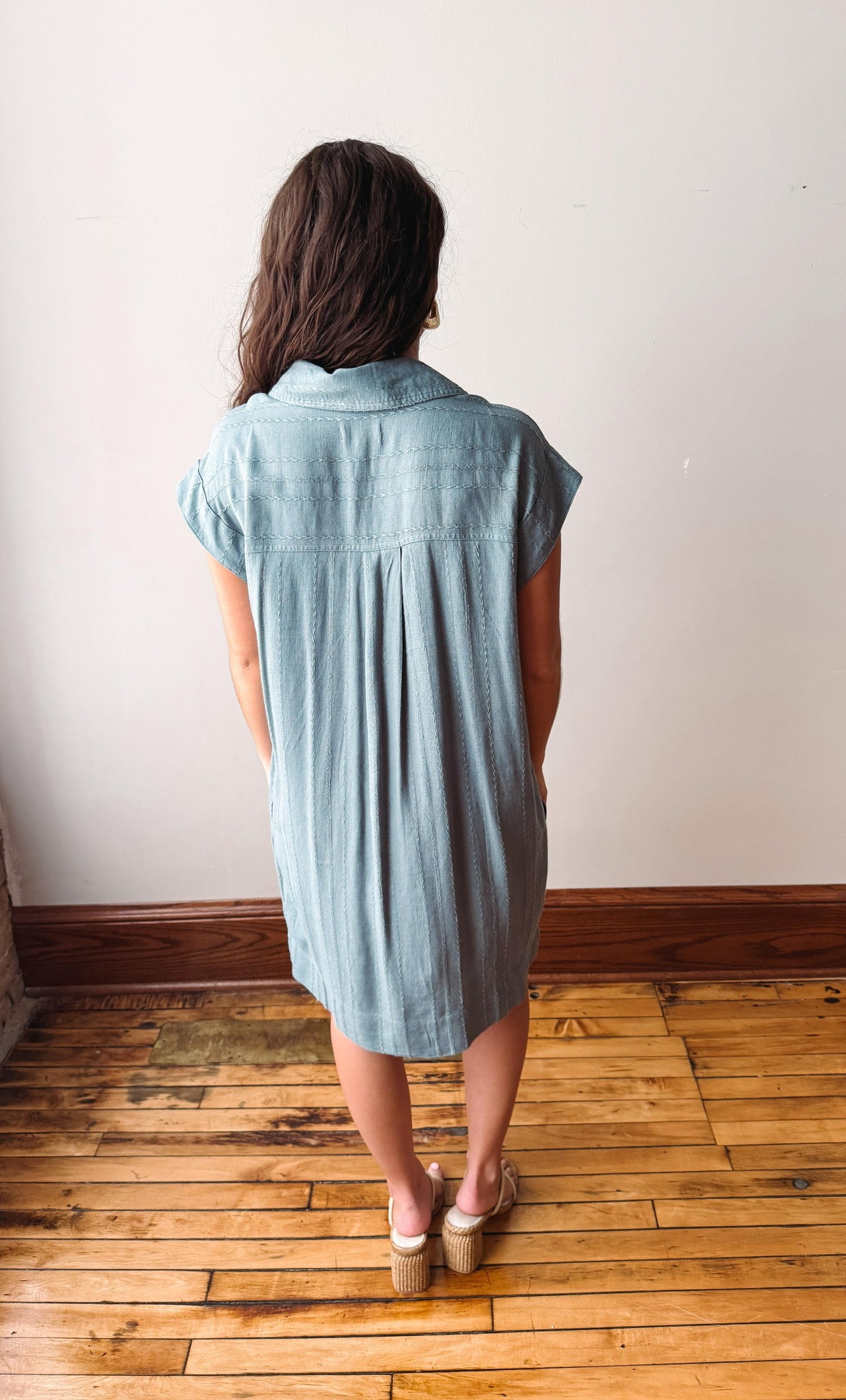 Woman wearing a light blue dress standing on a wooden floor with a white wall background