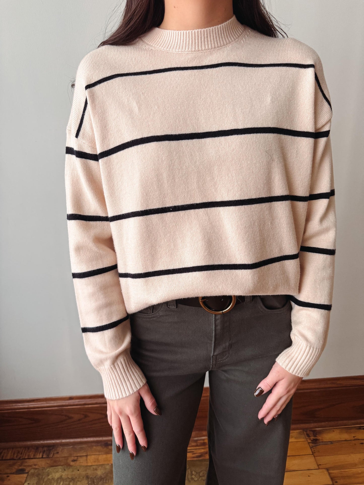 A person wearing a cream-colored knit sweater with horizontal black stripes, paired with dark gray pants, standing against a neutral indoor wall.