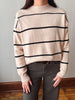A person wearing a cream-colored knit sweater with horizontal black stripes, paired with dark gray pants, standing against a neutral indoor wall.