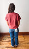 A person viewed from behind wearing a muted red short-sleeve t-shirt and blue jeans with a white tag attached to the back pocket, standing on a wooden floor against a plain white wall.