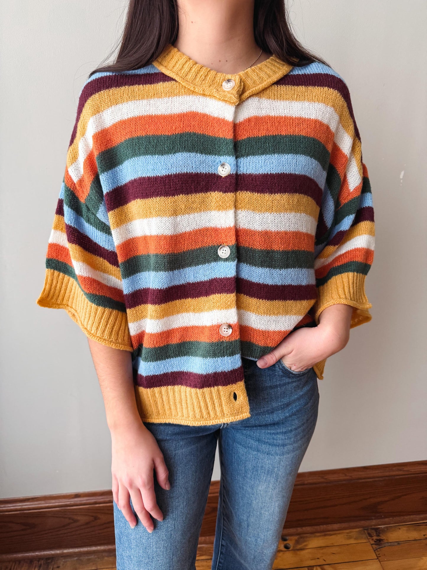 A person wearing a colorful striped knit cardigan sweater with mustard yellow trim, paired with blue jeans, standing indoors against a plain wall.