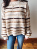 A person wearing a fuzzy knit sweater with horizontal beige, brown, and dark brown stripes, paired with blue jeans, standing indoors against a plain wall.