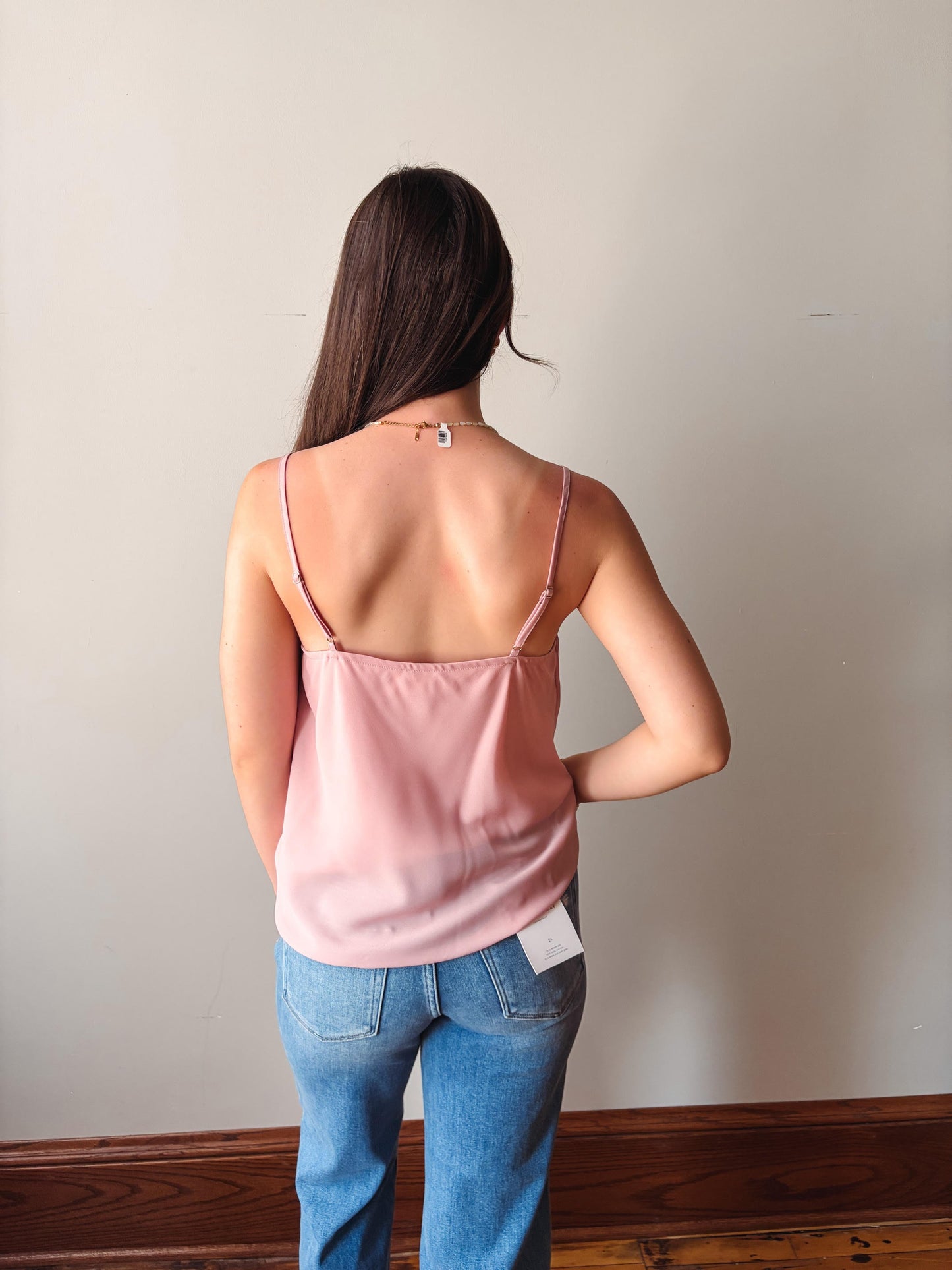 Woman wearing a pink tank top and blue jeans standing against a plain wall.