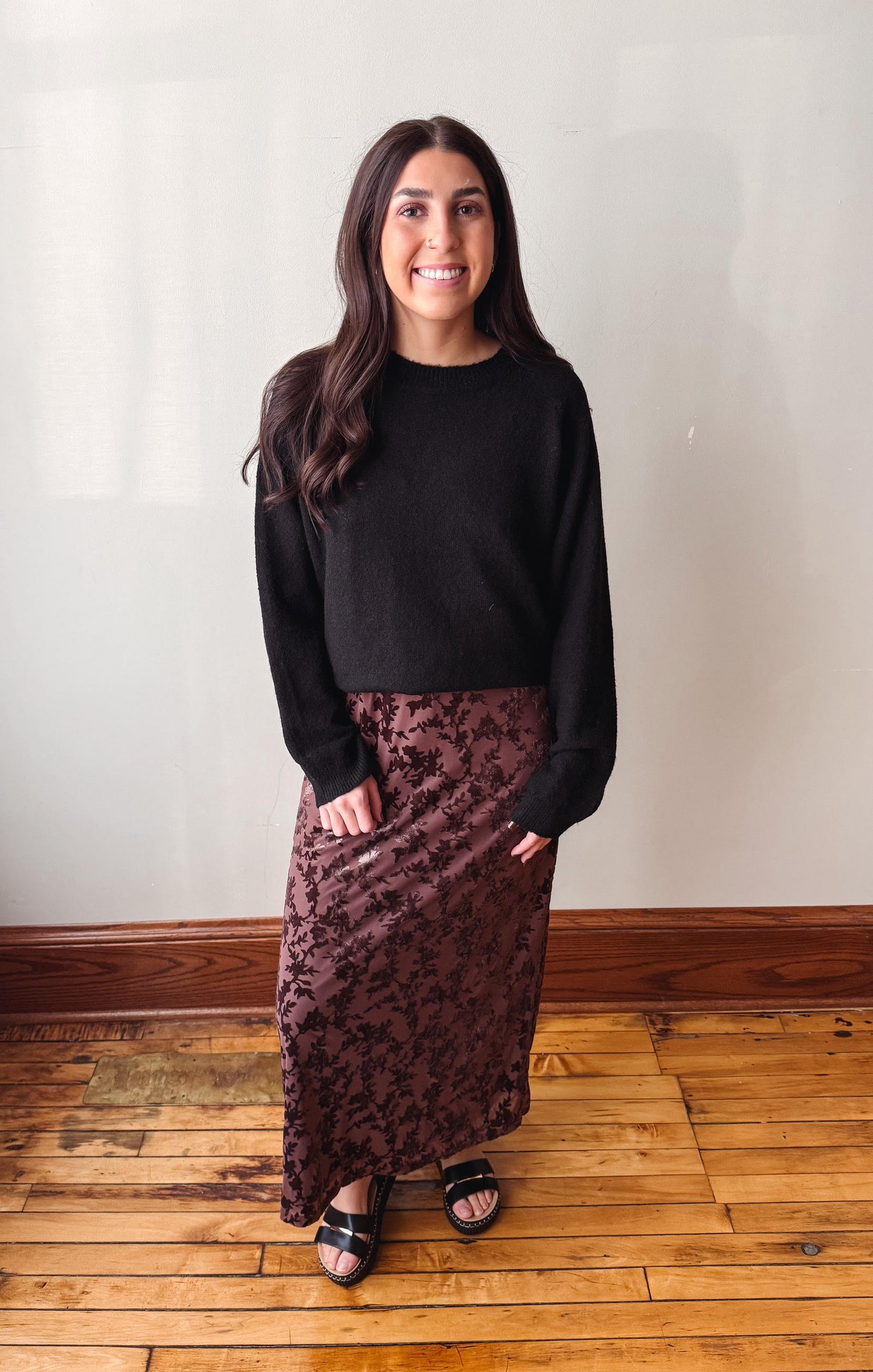 A woman stands indoors wearing a black long-sleeve knit top and a dark brown patterned midi skirt, paired with black slide sandals, against a plain off-white wall and wooden floor.