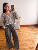 A woman taking a mirror selfie in a room with hardwood floors and a white wall, wearing a brown and cream striped cardigan and dark gray wide-leg pants.