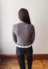 A woman viewed from behind wearing a gray knit sweater with a white hem detail and black jeans, standing on a wooden floor against a plain off-white wall.
