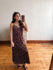 A woman takes a mirror selfie wearing a dark brown floral lace maxi dress with spaghetti straps and brown block-heel sandals in a room with light-colored walls and wooden floors.