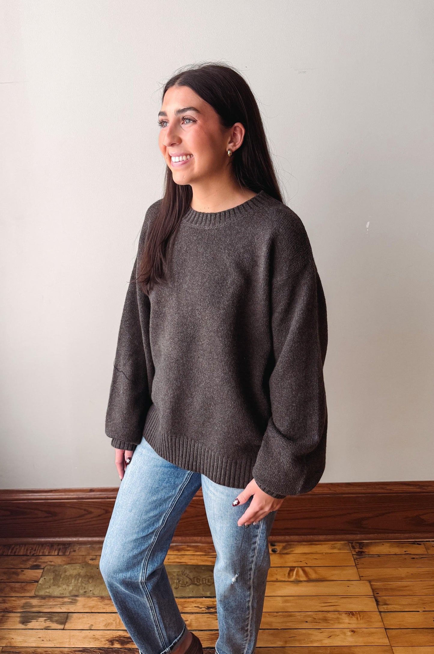 A woman wearing a dark brown knit crewneck sweater and light blue jeans, standing in a room with a light-colored wall and wooden floor.