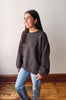 A woman wearing a dark brown knit crewneck sweater and light blue jeans, standing in a room with a light-colored wall and wooden floor.