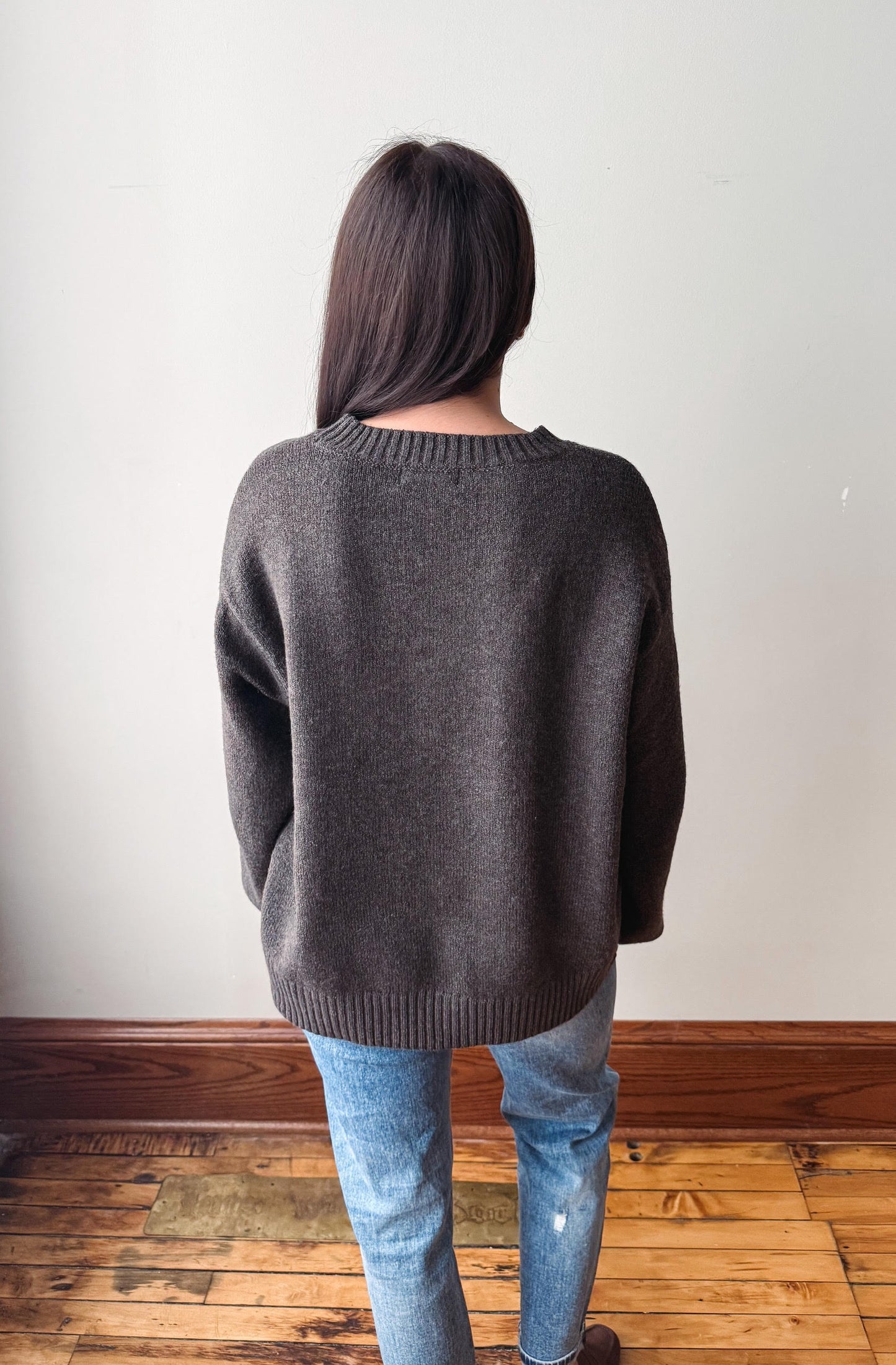 A person viewed from behind wearing an oversized dark gray knit sweater and light blue jeans, standing on a wooden floor against a plain off-white wall.