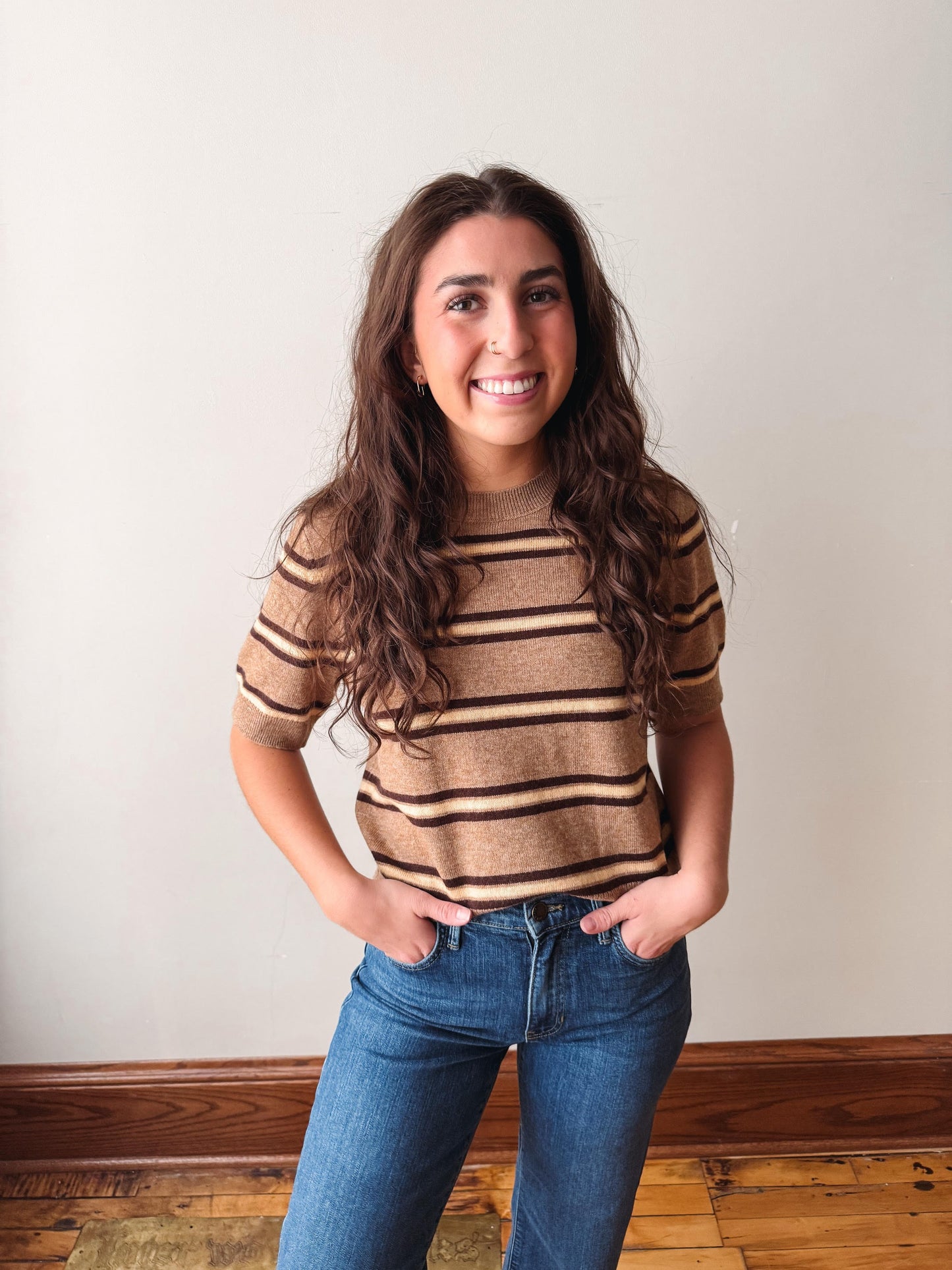 A smiling young woman with long wavy brown hair stands indoors wearing a brown and tan striped short-sleeve knit top and blue jeans against a plain off-white wall with wood trim and a wood floor.