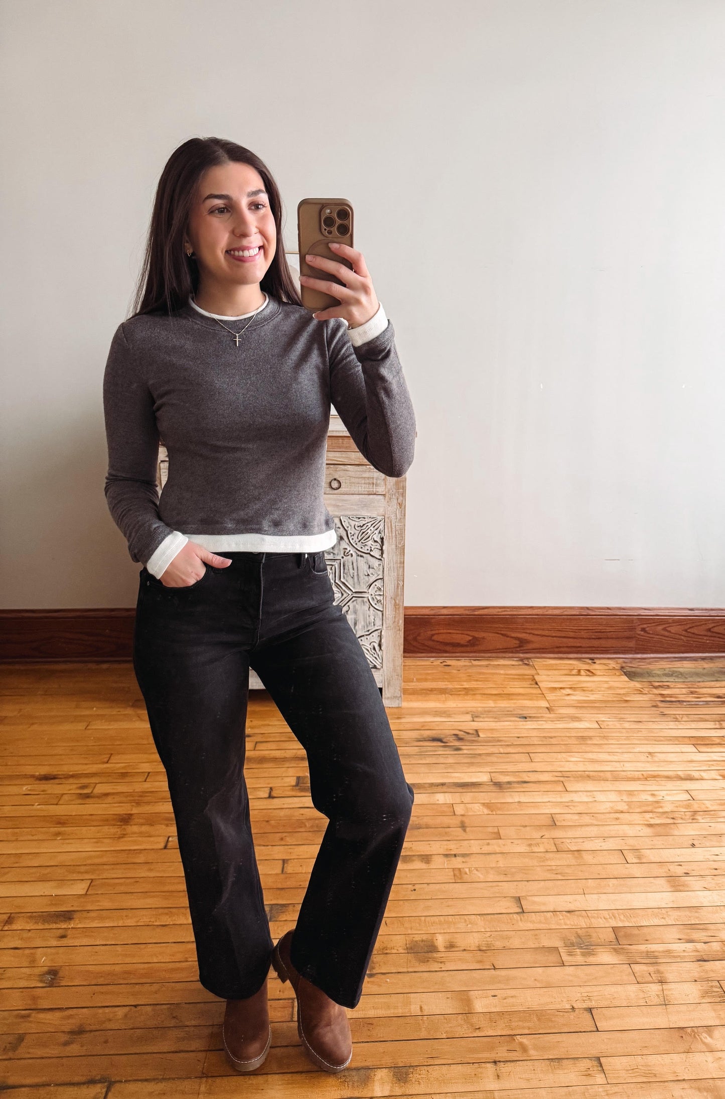 A woman takes a mirror selfie wearing a dark gray long-sleeve knit sweater with white ribbed cuffs and hem, black wide-leg jeans, and brown ankle boots, standing in a room with a light gray wall and wooden floor.