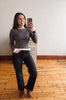 A woman takes a mirror selfie wearing a dark gray long-sleeve knit sweater with white ribbed cuffs and hem, black wide-leg jeans, and brown ankle boots, standing in a room with a light gray wall and wooden floor.