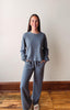 Woman wearing a gray sweater and pants standing against a plain wall.