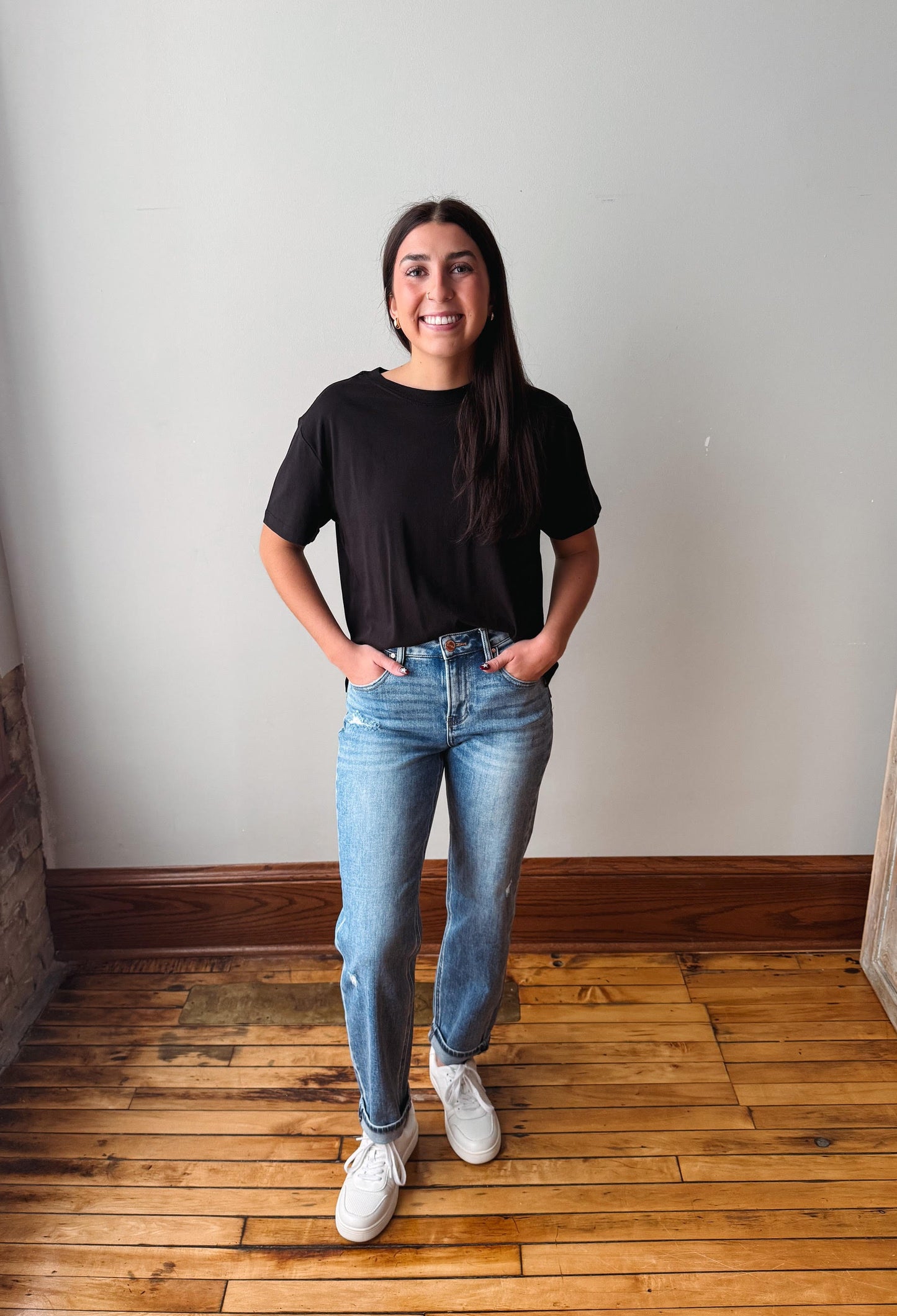 A woman stands in a room with a light gray wall and wooden floor, wearing a black short-sleeve t-shirt, light-wash blue jeans, and white sneakers.
