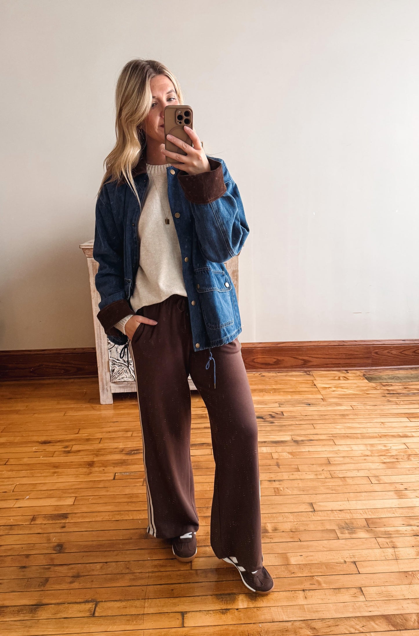 A woman takes a mirror selfie in a room with light wood floors and a white wall, wearing a blue denim jacket with brown ribbed trim over a cream knit top and dark brown wide-leg sweatpants.