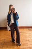 A woman takes a mirror selfie in a room with light wood floors and a white wall, wearing a blue denim jacket with brown ribbed trim over a cream knit top and dark brown wide-leg sweatpants.
