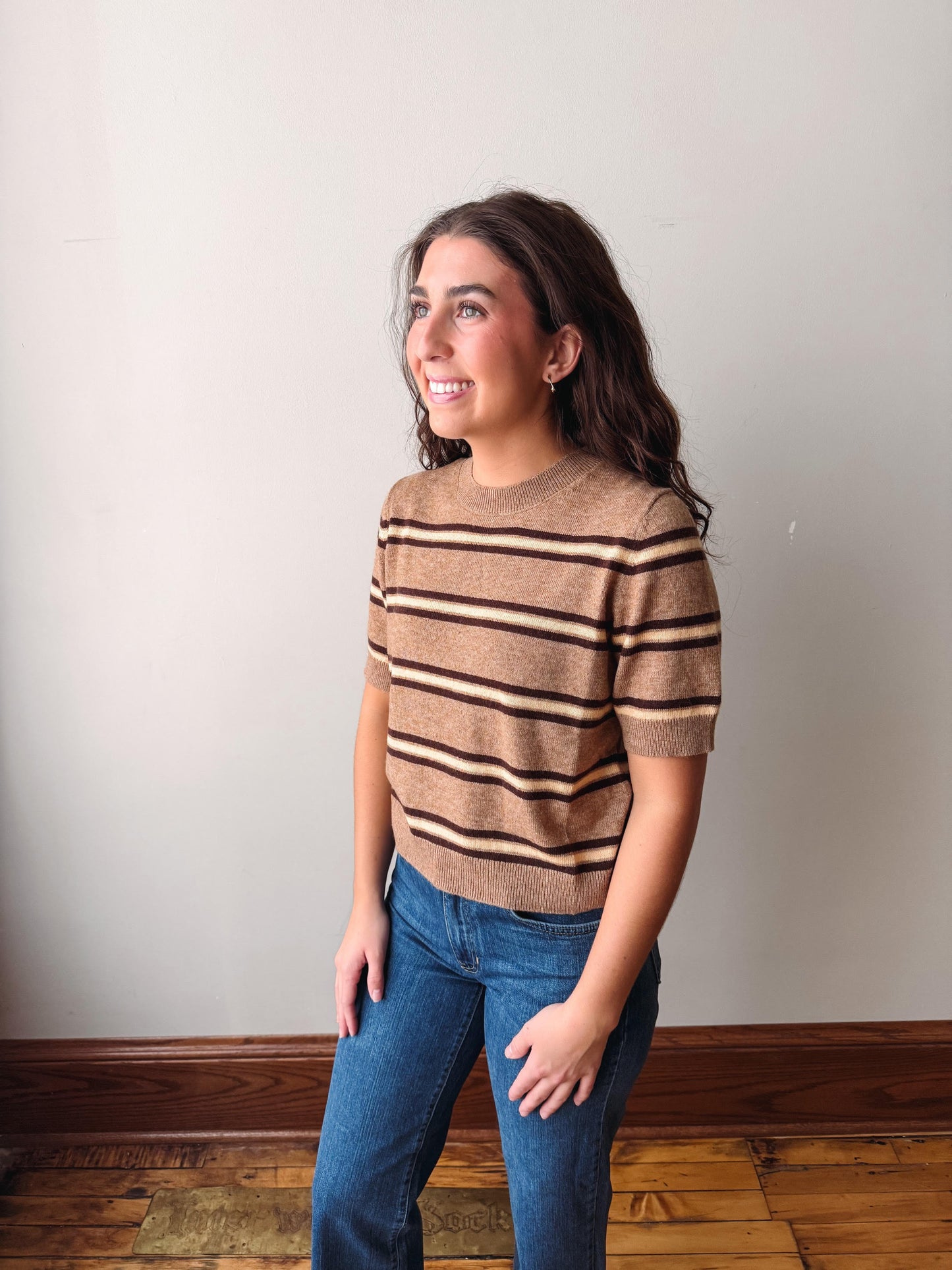 A woman modeling a short-sleeve knit sweater with horizontal brown and beige stripes, paired with blue jeans, standing against a plain off-white wall with a wooden floor.