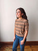 A woman modeling a short-sleeve knit sweater with horizontal brown and beige stripes, paired with blue jeans, standing against a plain off-white wall with a wooden floor.