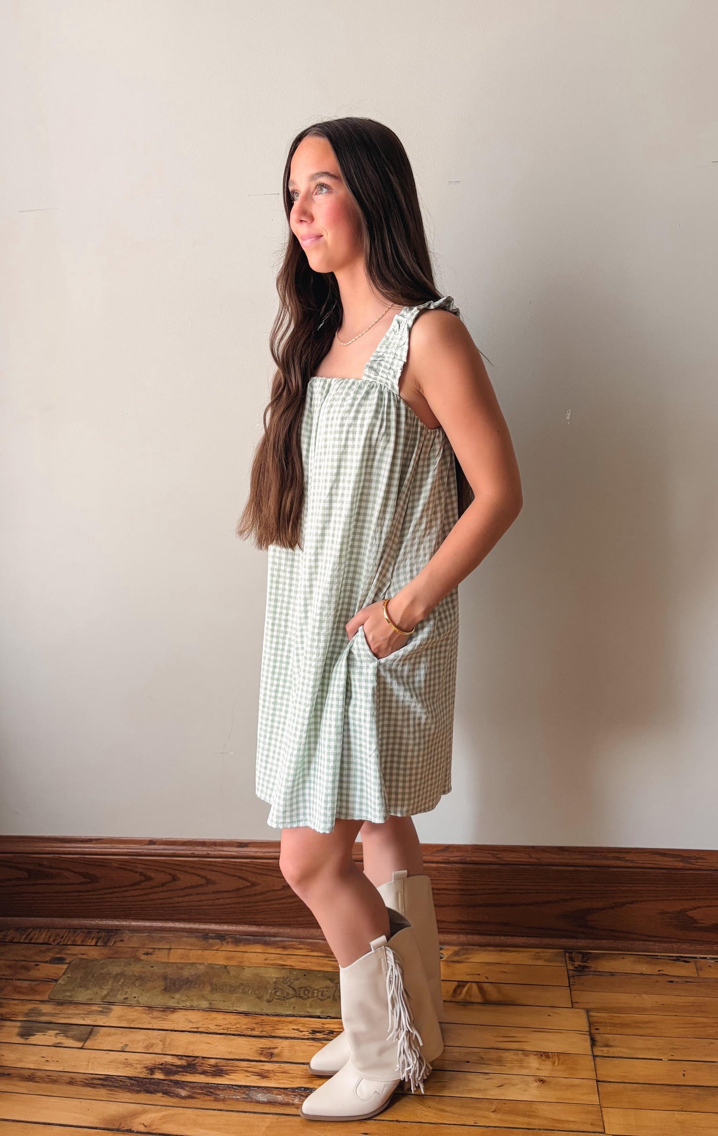 Woman wearing a green checkered dress and white boots standing against a plain wall.
