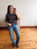 A woman takes a mirror selfie in a room with light wood floors and a white wall, wearing a brown and navy striped t-shirt, blue jeans, and brown leather boots.