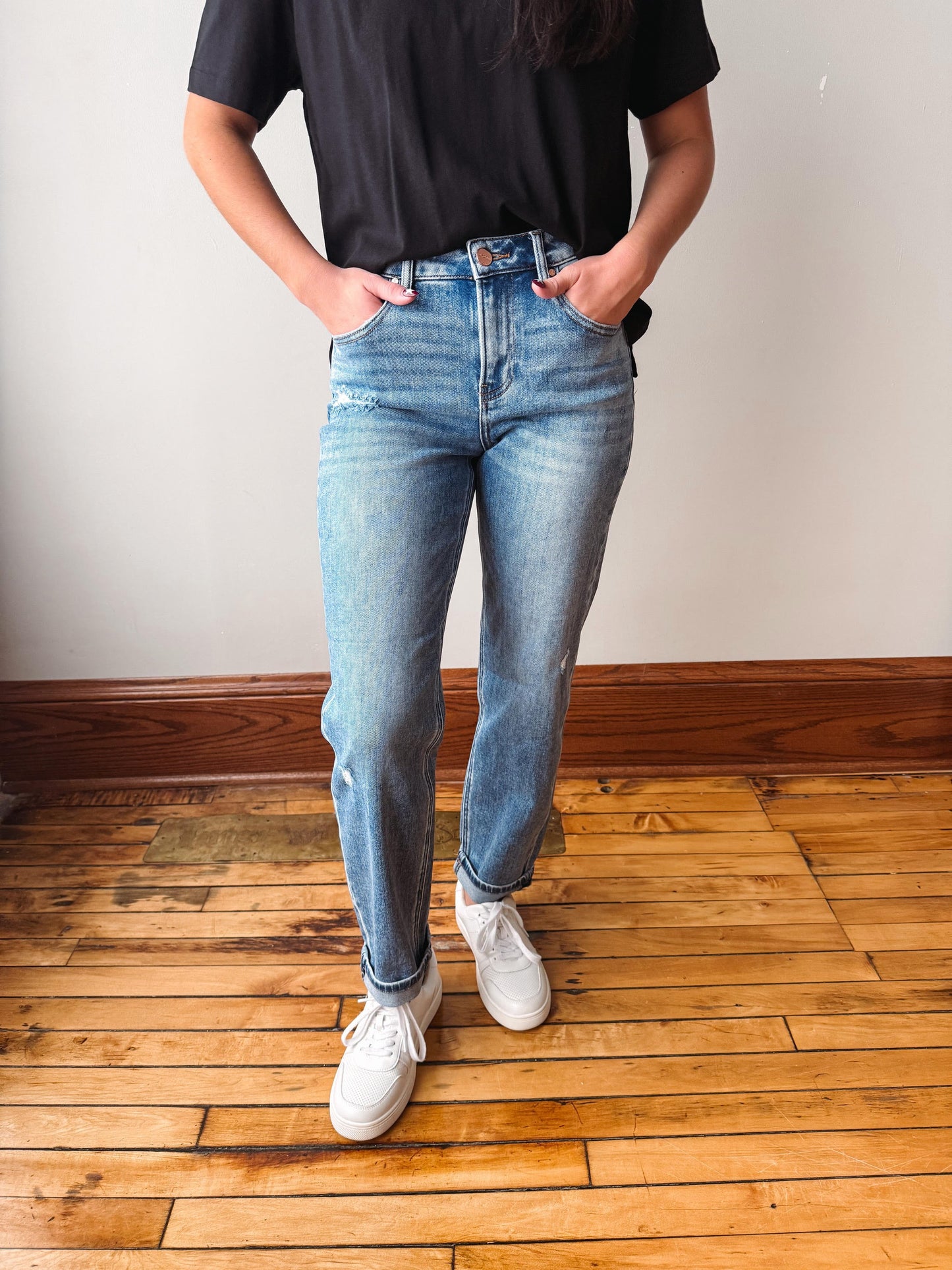 A person wearing light-wash blue jeans, a black t-shirt, and white sneakers, standing on a wooden floor against a plain wall.