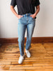 A person wearing light-wash blue jeans, a black t-shirt, and white sneakers, standing on a wooden floor against a plain wall.