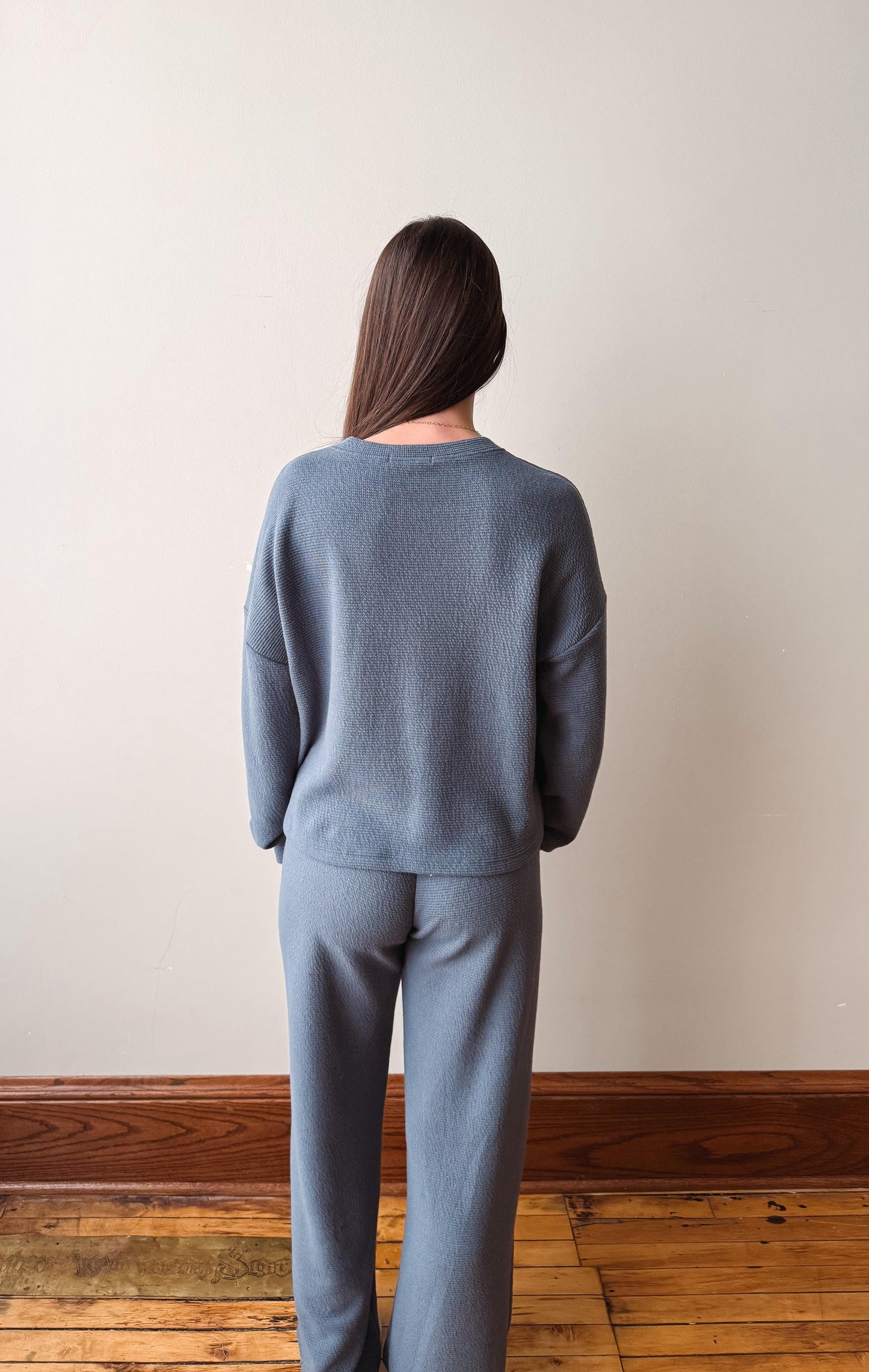 Person wearing a gray sweater and pants standing against a plain wall.