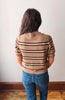A person viewed from behind wearing a beige short-sleeve knit sweater with dark brown and cream horizontal stripes, paired with blue jeans, standing against a plain off-white wall.