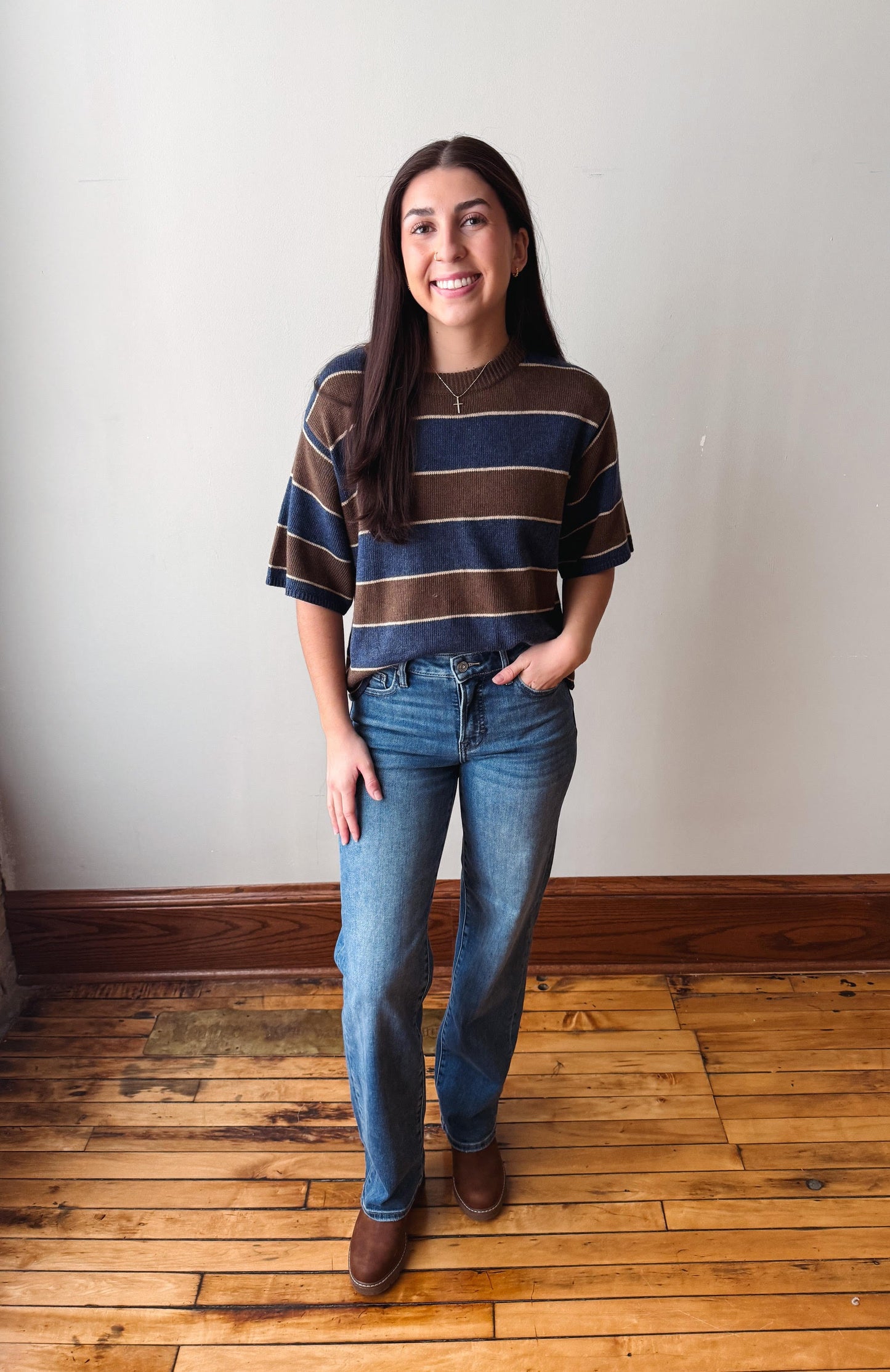 A woman stands in a room with a white wall and wooden floor, wearing a brown and navy blue striped short-sleeve t-shirt, blue jeans, and brown boots.