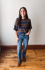 A woman stands in a room with a white wall and wooden floor, wearing a brown and navy blue striped short-sleeve t-shirt, blue jeans, and brown boots.
