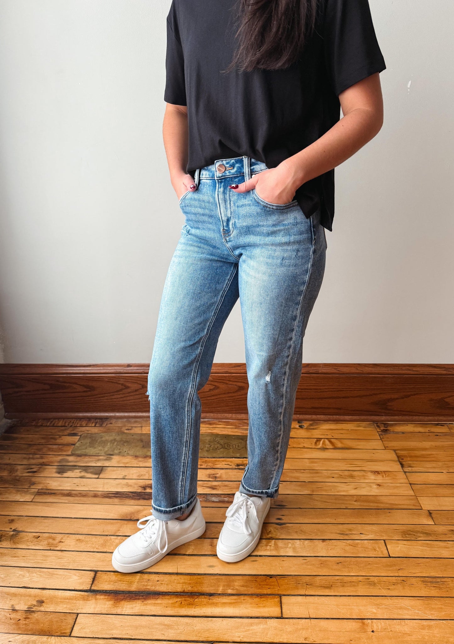 A person wearing light-wash blue jeans, a black t-shirt, and white sneakers, standing on a wooden floor against a plain wall.