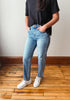 A person wearing light-wash blue jeans, a black t-shirt, and white sneakers, standing on a wooden floor against a plain wall.