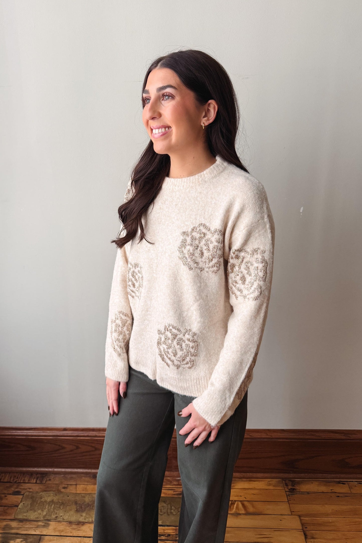 A woman wearing a cream-colored knit sweater with textured floral patterns on the front and sleeves, paired with dark gray pants, standing in a room with a light gray wall and wooden floor.