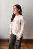 A woman wearing a cream-colored knit sweater with textured floral patterns on the front and sleeves, paired with dark gray pants, standing in a room with a light gray wall and wooden floor.