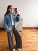 A woman taking a mirror selfie in a room with hardwood floors and a white wall, wearing a light blue cropped denim jacket over a white t-shirt and wide-leg dark gray pants.