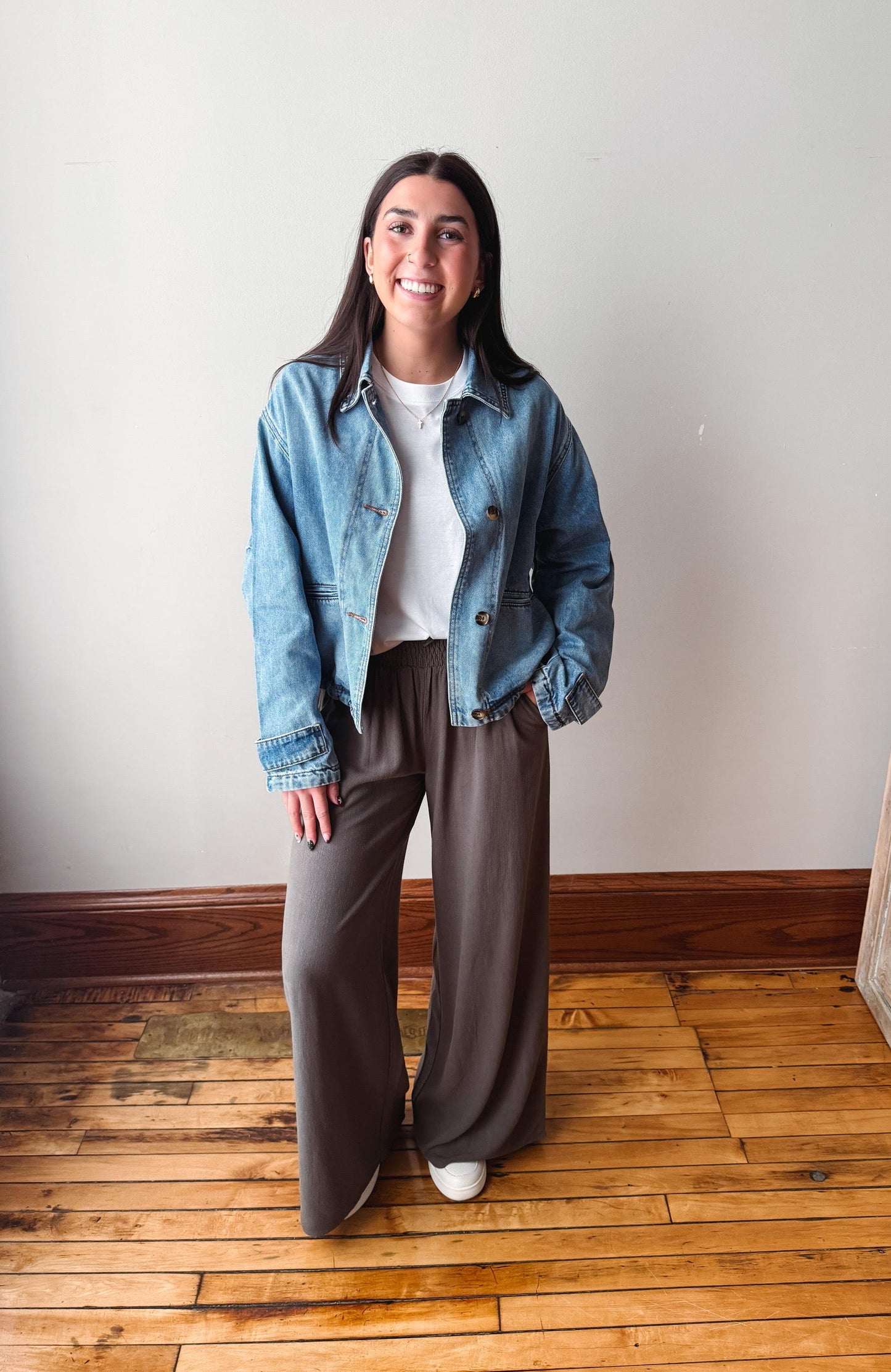 A woman stands in a room with a white wall and wooden floor, wearing a light blue denim jacket over a white t-shirt and brown wide-leg pants.
